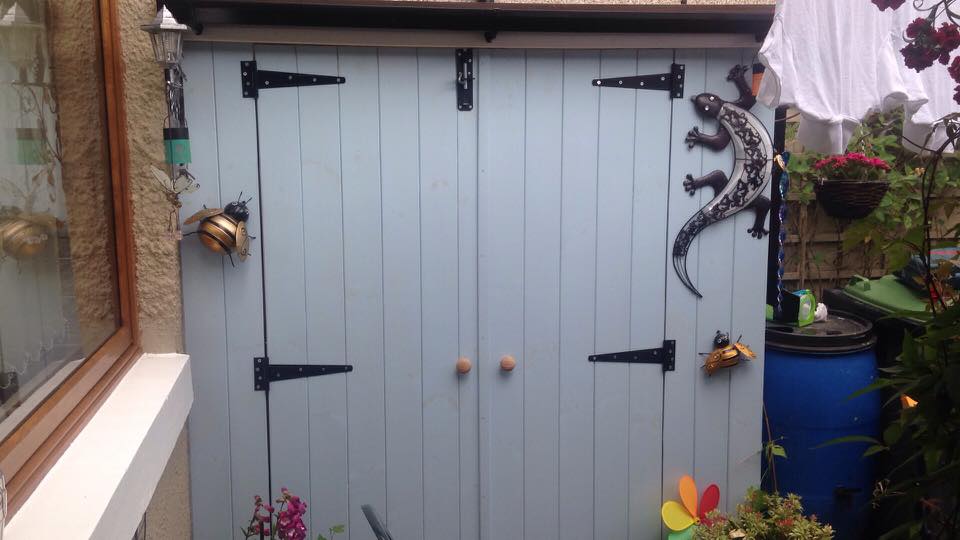 A shed painted pale grey with black T-hinges, garden ornaments around it
